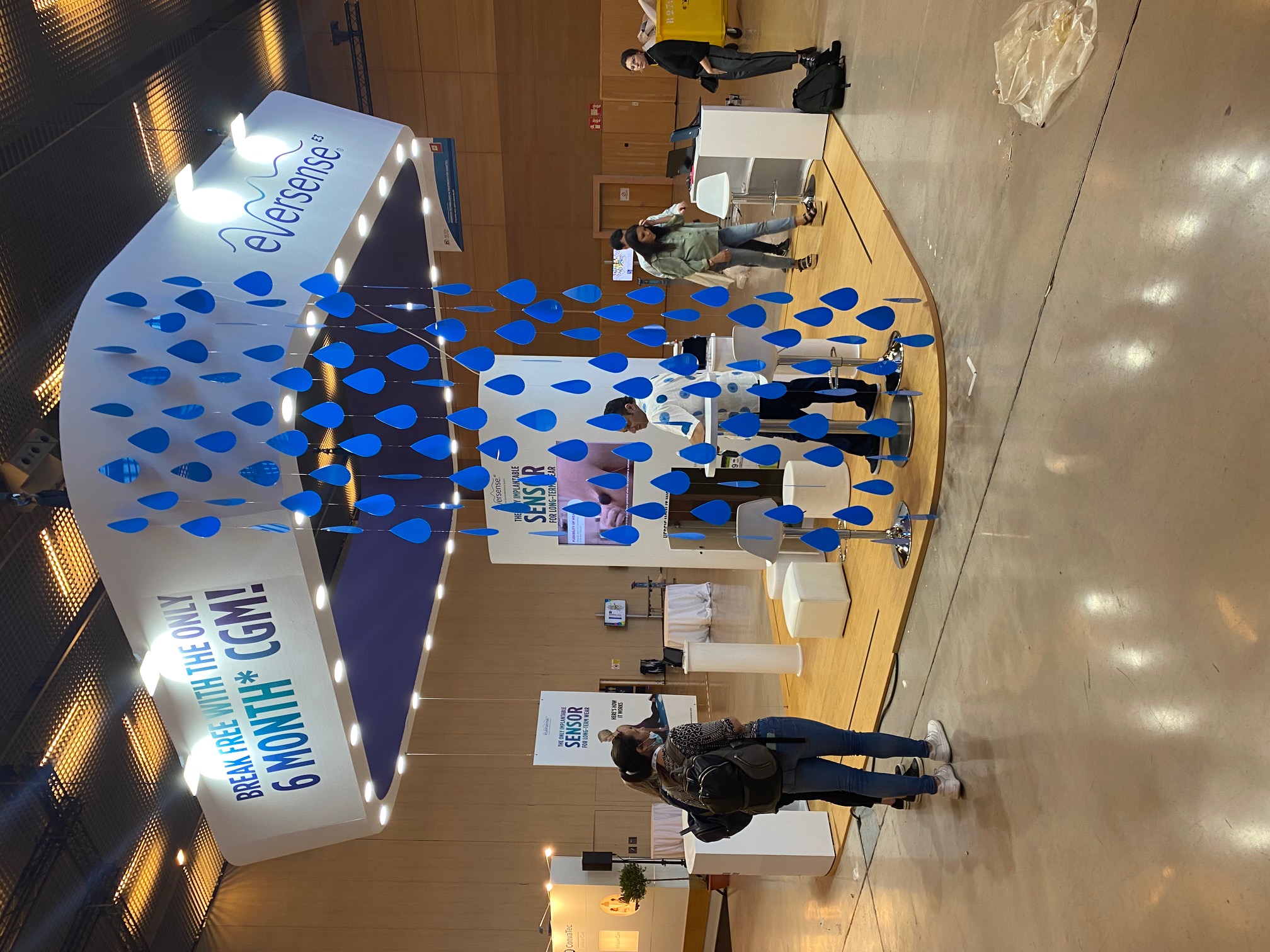 Stand de Eversense CGM en congreso médico con estructura curva blanca, gotas azules decorativas colgantes y gráficas de gran formato